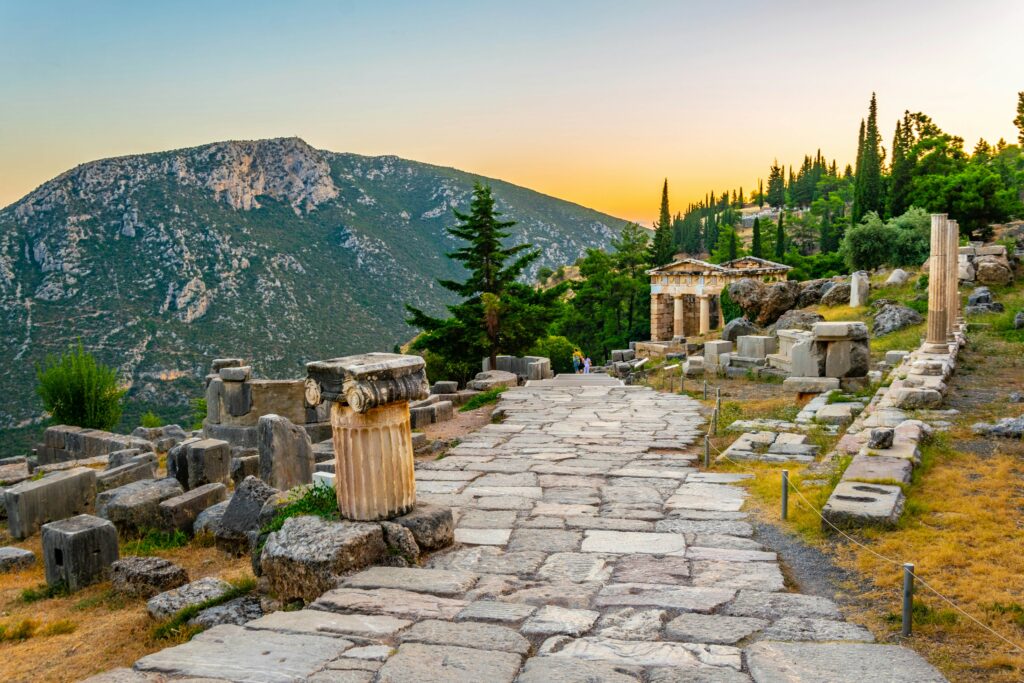 Αρχαία πέτρινη οδός στον αρχαιολογικό χώρο των Δελφών με στήλες και βουνό στο φόντο.