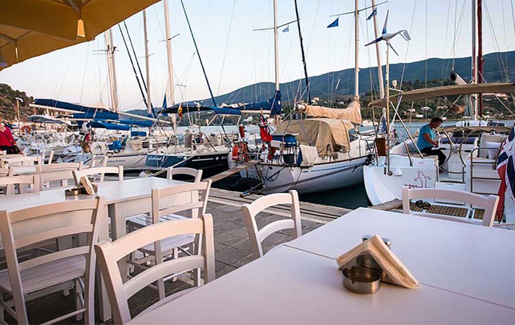 Τραπεζάκια εστιατορίου στον Πόρο δίπλα σε ιστιοπλοϊκά και θέα στο λιμάνι.