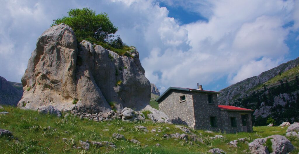 Ορειβατικό καταφύγιο στη Γκιώνα κάτω από έναν τεράστιο βράχο, μέσα στο πράσινο τοπίο του καλοκαιριού