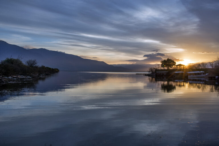Τεχνητή Λίμνη Κερκίνης – Ηλιοβασίλεμα στη λίμνη με ήρεμα νερά και φόντο τα βουνά Μπέλες